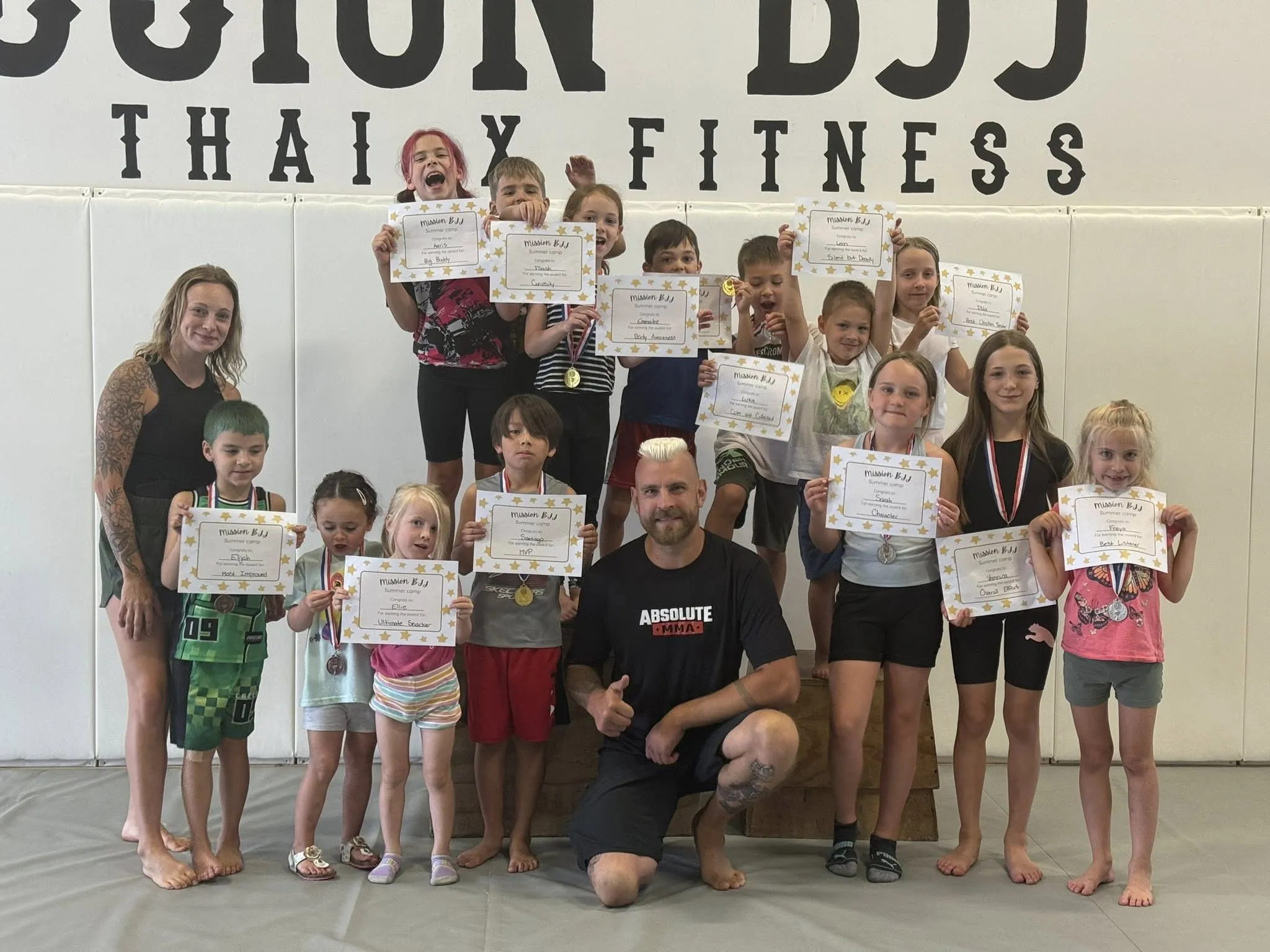 Kids celebrating with their summer camp certificates alongside coaches at Mission BJJ